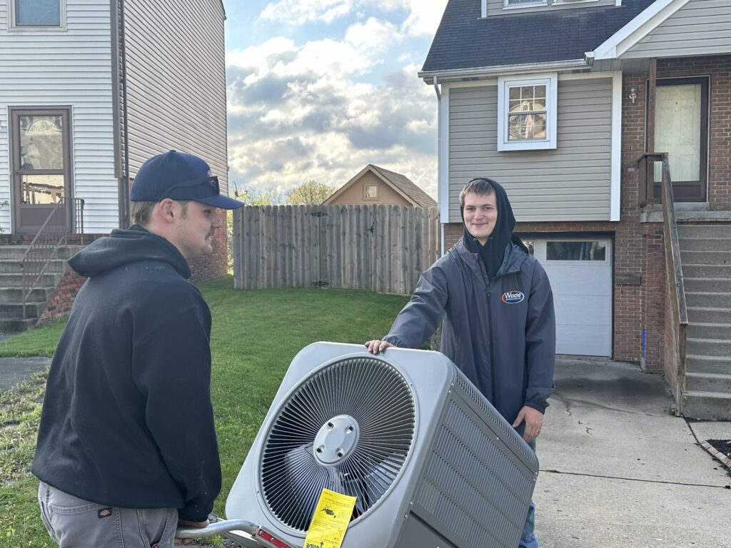 Two Wade Heating & Cooling technicians wheeling an AC condenser unit onto a property for installation